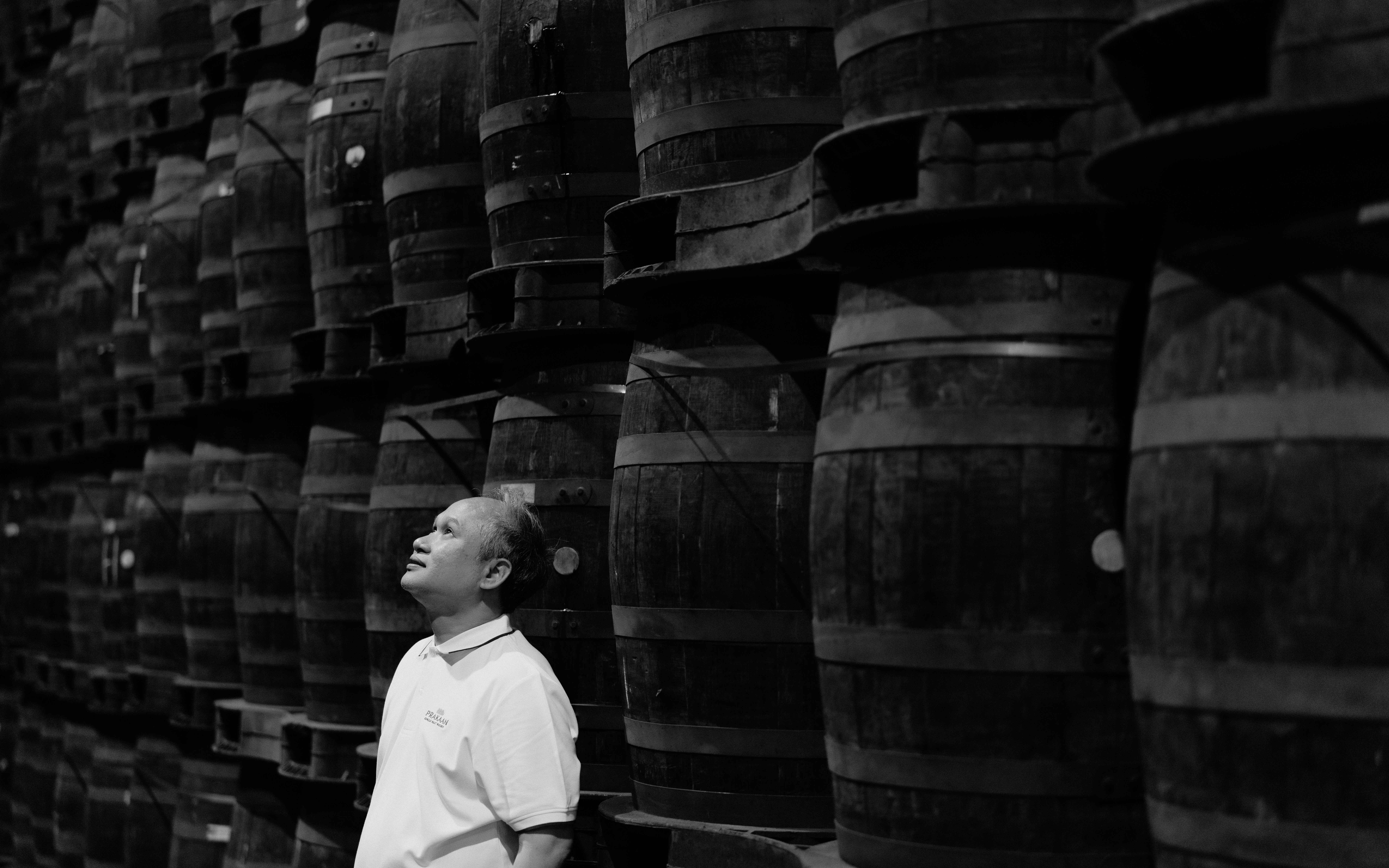 A black and white photo of a man standing with his back to a stack of whisky casks